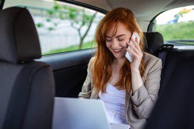 Dizüstü bilgisayarda mutlu kızıl saçlı kadın ve araba arka koltuğunda telefon. Gülümseyen kadın girişimci takside bilgisayar ve akıllı telefonla birden fazla iş yapıyor. Uzak çalışma, başarı ve iş iletişimi kavramı.