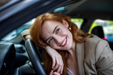 Direksiyona yaslanan ve kameraya gülümseyen mutlu, genç, kızıl saçlı kadın. Güzel bayan sürücü araçla yolculuğun tadını çıkarıyor. Otomobil yaşam tarzı, ulaşım ve araba sigortası kavramı.
