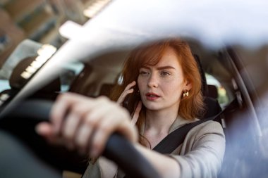 Stresli genç, kızıl saçlı iş kadını, yoğun bir şehirde araba kullanırken akıllı telefondan konuşuyor. Profesyonel bronz bir ceket ve modern bir yerleşim bölgesinde altın küpeler takıyor.