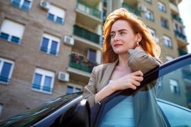 Güzel kızıl bir kadın, modern bir şehir mahallesinde, altın saat boyunca arka planda apartman binaları olan bir şehir ortamında arabasının kapısına yaslanmış, düşünceli bir şekilde yüzünü çeviriyor.
