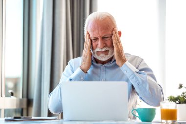 Başağrısı olan stresli son sınıf öğrencisi dizüstü bilgisayarda çalışıyor, Olgun Erkek Duyguları Uzaktan Çalışmanın etkisi altında. Bilgisayar ve iş çizelgeleriyle masada otururken başını ağrıtan yaşlı kişi.
