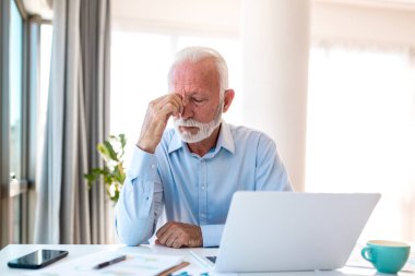 Dijital Göz Gerilimi 'nden yorgun yaşlı adam gözlerini ovuşturuyor, dizüstü bilgisayarda çalışırken yorgun hisseden olgun erkek. Parlak ev ofisinde bulanık görme ve yorgunluk çeken yaşlı bir profesyonel..