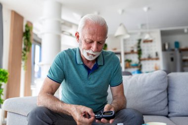 Kıdemli adam kan şekeri seviyesini glikoz sayacıyla ölçüyor. Şeker hastalığı olan yaşlı bir erkek hasta evde insülin testi yapıyor. Sağlık ve tıp konsepti. Kanepede dijital glukometre kullanan kişi.