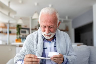 Hasta kıdemli adam evde termometreyi kontrol ediyor. Grip veya soğuk algınlığı semptomları olan yaşlı bir erkek hasta battaniyeyle kaplı kanepede oturuyor. Sağlık ve tıp konsepti. Yüksek vücut sıcaklığı.