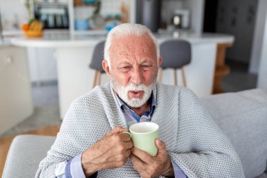 Hasta kıdemli adam kanepede sıcak çay içiyor. Soğuk algınlığı ya da grip belirtileri gösteren yaşlı bir erkek hasta. Sağlık ve tıp konsepti. Elinde bir kupa sıcak bitkisel içecek tutan kişi..
