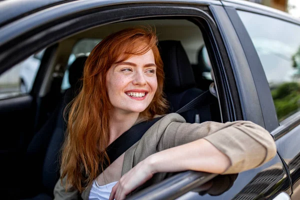 Neşeli kızıl saçlı kadın, güneşli bir sabah güneşli bir mahallede arabasının içinden yüzünü çeviriyor ve gülümsüyor. Arka planda apartmanlar başarılı bir yaşam tarzı gösteriyor.