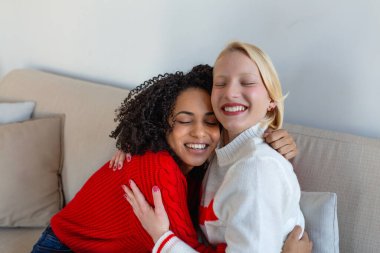 Mutlu çeşitli bayan arkadaşlar evdeki kanepede kucaklaşıyorlar. Çoklu etnik arkadaşlık, destek ve aşk konsepti. Gözleri kapalı, gülümseyen, kırmızı beyaz süveter giyen iki genç kadın..