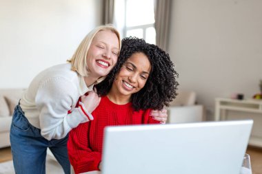 Evde dizüstü bilgisayar kullanırken sarılıp gülen mutlu genç kadınlar. Çeşitli Kadın Arkadaşlığı, Bağlanma ve Şefkat Konsepti. Neşeli Kızlar Oturma Odasında Not Defteri 'nde Komik Videolar İzliyor