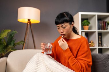 İlaç alıyorum. Kadın hap yutuyor, sağlık ve eczane konsepti evde tıbbi tedavi görüyor. Turuncu kazaklı hasta Japon kadın bir bardak su ve kapsülle kanepede oturuyor..
