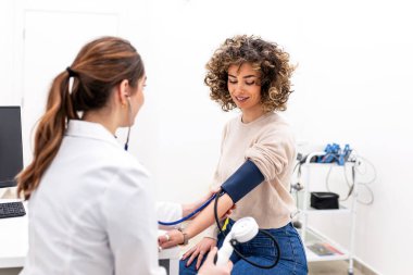Gülümseyen kadın doktor klinikteki kıvırcık saçlı kadının kan basıncını ölçüyor. Profesyonel doktor hastanın kalp sağlığını kontrol ediyor. Yetişkin hastalarda sağlık ve tıp konsepti.
