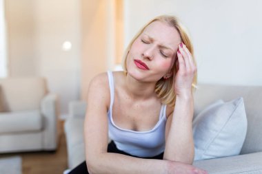 Migren ve küme baş ağrısı. Tapınak ağrısı ve stresi olan kadın, Sağlık ve Tıp konsepti. Çekici sarışın kız kanepeye oturmuş, gözleri kapalı, acı dolu kafasını tutuyor..