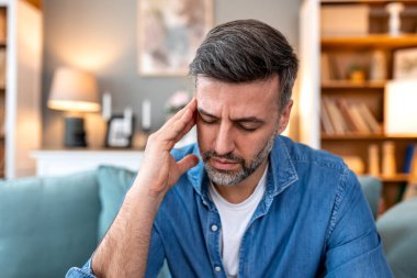 Baş ağrısı ve migren. Kanepede eli ve gözleri kapalı, acı dolu bir tapınağı tutan yorgun bir adam, Sağlık ve Tıp konsepti. Orta yaşlı erkek hasta ve evde baş ağrısı çekiyor..