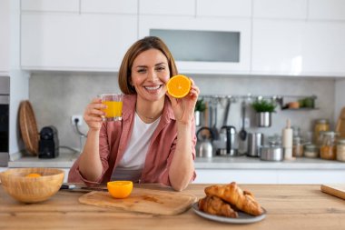 C vitamini ve sağlıklı kahvaltı. Elinde bir bardak portakal suyu ve taze portakal suyu tutan gülümseyen kadın sağlık sigortası ve ilaç konsepti. Mutfakta kruvasan ve meyvelerle oturan mutlu bir kadın..