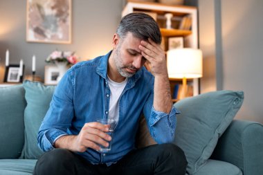 Baş ağrısı ve migren. Elinde bir bardak su olan bir adam kanepeye oturmuş, eli, sağlık sigortası ve ilaç konseptiyle acı dolu bir kafa tutuyor. Orta yaşlı bir adam kendini hasta hissediyor ve evinde baş ağrısı çekiyor..