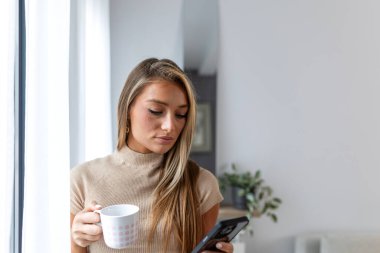 Pencere kenarında elinde kahve fincanı tutan ve akıllı telefon kullanan genç bir kadın. Kadın sosyal medyayı kontrol ediyor ya da evde cep telefonuyla mesajlaşıyor. Teknoloji, günlük rutin ve rahatlama kavramı.