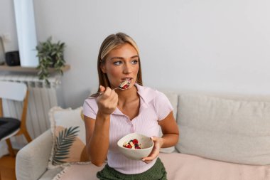 Kanepede sağlıklı kahvaltı yapmak. Meyveli ve muzlu mısır gevreğinin tadını çıkaran genç bir kadın. Sağlık ve beslenme konsepti. Çekici bir kadın kanepede oturup organik sabah yemeği yiyor..