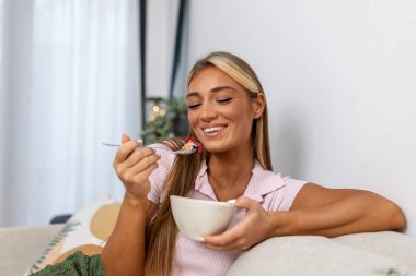 Evde sağlıklı bir kahvaltı. Kase, Sağlık ve Beslenme konsepti böğürtlenli gevrek yiyen mutlu kadın. Çekici sarışın kız kanepede oturuyor ve gülümseyerek lezzetli bir sabah yemeğinin tadını çıkarıyor..