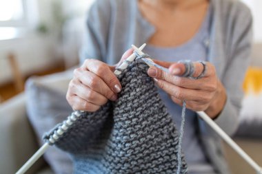 Gri yün ve beyaz iğnelerle örgü ören yaşlı kadınların ellerini kapatın. El işi, manuel terapi ve yaratıcı bir hobi. Olgun bir insan evde kanepede örgü örmeye çalışıyor..