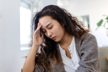 Baş ağrısı ve migren. Akıl sağlığı, stres sorunu olan insanlar, sağlık sigortası ve tıp konsepti. Kanepede oturan yorgun Asyalı kadın ve elinde ağrılı bir kafayla evde oturuyor. Yorgun bir kadın..