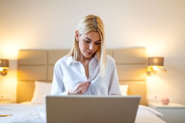Otelde dizüstü bilgisayarda çalışan profesyonel bir kadın. Beyaz gömlekli güzel sarışın iş kadını iş seyahatleri sırasında yatakta oturup bilgisayar kullanıyor. Yönetici yaşam tarzı ve konukseverlik kavramı.