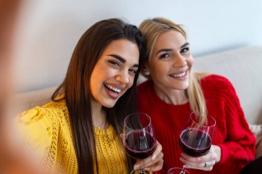 Mutlu genç kadınlar kırmızı şarap içerken selfie çekiyorlar. İki güzel bayan arkadaş evde şarap kadehleriyle gülüp eğleniyorlar. Kutlama ve yaşam tarzı konsepti. Gülümseyen neşeli kızlar.