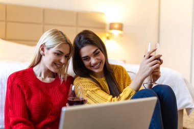 Laptop kullanan ve yatak odasında kırmızı şarap içen iki genç bayan arkadaş. Mutlu kadınlar yerde otururken bilgisayarda film izliyorlar. Teknoloji ve eğlence kavramı. Güzel kızlar eğleniyor..