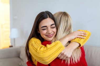 Mutlu genç bir kadın evde arkadaşına sarılıyor. Gülümseyen bir kadın oturma odasında en iyi arkadaşını kucaklıyor. Arkadaşlık, destek ve yaşam tarzı konsepti. Renkli kazaklı iki kız şefkat gösteriyor..