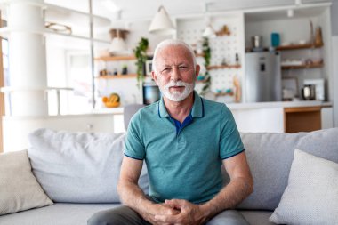 Parlak modern oturma odasındaki koltukta oturan gülümseyen son sınıf öğrencisinin portresi. Mutlu yaşlı insan evde emeklilik hayatı yaşıyor. Yaşlanma, sağlık ve ev hayatı kavramı. Ön görünüm.