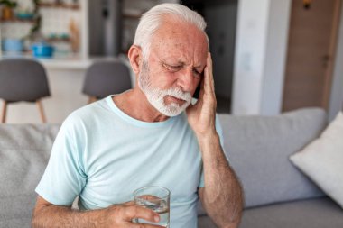 Baş ağrısı olan yaşlı bir adam bir bardak su tutuyor ve kanepede şakağına dokunuyor. Migren, hipertansiyon ya da susuzluk çeken yaşlı bir insan. Sağlık ve tıp konsepti. Ağrı yönetimi.
