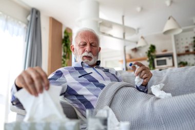 Hasta kıdemli adam termometre ve mendillerle kanepede yatıyor, ateşi kontrol ediyor. Grip, soğuk algınlığı ya da virüs enfeksiyonu belirtileri gösteren yaşlı bir insan. Sağlık ve tıp konsepti. Evde tedavi ve hemşirelik..