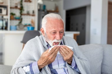 Hasta kıdemli adam mendile hapşırıyor ve kanepeye battaniye örtüyor. Mevsimsel grip, soğuk algınlığı ya da alerji belirtileri gösteren yaşlı bir insan. Sağlık ve tıp konsepti. Emeklilik ve sağlık sorunları.