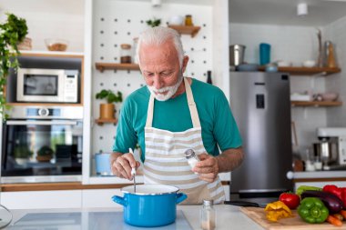 Kıdemli adam mutfakta sağlıklı sebze çorbası pişirirken tencereye tuz ekliyor. Yaşlı kişi apronla besleyici yemek hazırlıyor. Sağlıklı yaşam tarzı, ev yemekleri ve emeklilik hobisi.