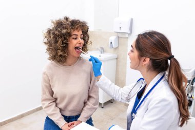 Kadın doktor kadın hastaya tahta spatulayla boğaz muayenesi yapıyor. Klinikte bademcikleri kontrol eden bir doktor. Sağlık ve tıp konsepti. Tıbbi danışmanlık ve profesyonel kontrol.