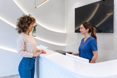 Klinik resepsiyonundaki hasta. Hastane resepsiyonu, sağlık ve tıp konsepti. Çekici bir kadın beyaz modern resepsiyonda dost canlısı bir doktorla konuşuyor. Profesyonel tıbbi servis..