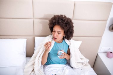 Hasta kadın yatakta hap alıyor. Battaniyeye sarılı, elinde su ve ilaç bardağı, grip ve ateş konsepti olan Afrika kökenli Amerikalı bir kadın. Genç bayan kendini iyi hissetmiyor ve evde üşütüyor..