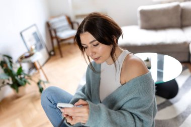 Evde çalışırken akıllı telefon kullanan bir kadının yüksek açılı görüntüsü. Genç bayan profesyonel mesajları kontrol ediyor ya da modern oturma odasındaki mobil uygulama. Dijital göçebe yaşam tarzı kavramı.