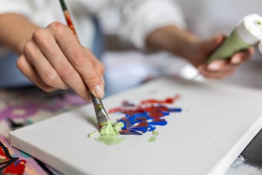 Beyaz bir tuvalde yeşil ve mavi akrilik boyayı fırçayla karıştıran bir sanatçının detaylı görüntüsü. Parlak minimalist yaratıcı çalışma alanında dokulu soyut sanat üzerine çalışan profesyonel kadın yaratıcı.