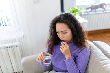 Genç Asyalı kadın evinde bir bardak suyla ilaç alıyor. Sağlık ve tıp konsepti. Hasta kız battaniyenin altında kanepede oturup grip için antibiyotik, ağrı kesici ya da soğuk algınlığı ilacı alıyor..