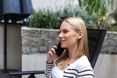 Açık havada akıllı telefondan konuşan mutlu bir kadın. İletişim ve yaşam tarzı konsepti. Bahçede ya da terasta oturan genç bir bayan cep telefonuyla arıyor. Gülümseyen kız cep telefonu kullanıyor..