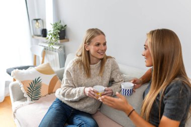 Kanepede kahve fincanlarıyla konuşan iki bayan arkadaş. İletişim, dostluk ve eğlence konsepti. Oturma odasında sohbet eden mutlu genç kadınlar. Ev hayatı, paylaşım ve ev sohbeti.