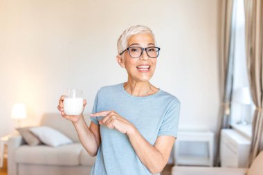 Gözlüklü ve bir bardak sütü işaret eden mutlu son sınıf kadını. Olgun bir bayan, kemik sağlığı için sağlıklı içeceği ve kalsiyumu teşvik ediyor. Sağlık ve tıp konsepti. Evdeki yaşlı kişi..
