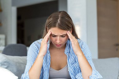 Güçlü baş ağrısı ve migrenden muzdarip genç bir kadın. Sağlık ve tıp konsepti. Stresli insan koltukta oturur ve elleriyle başını tutar. Yorgun, yorgun ya da bunalmış hisseder..