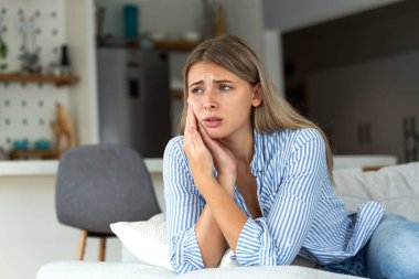 Diş ağrısı ve diş problemi kavramı. Sağlık ve ilaç servisi. Üzgün genç kadın kanepede oturuyor ve elleriyle acı dolu yanağını tutuyor, güçlü diş ağrısı, hassas dişler ya da çürüme yüzünden acı çekiyor..