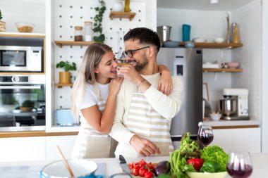 Neşeli çift mutfakta yemek pişirirken yemeği tadıyor. Sağlıklı beslenme ve romantik ilişki kavramı. Erkek kadını taze vişneli domatesle beslerken evde birlikte sağlıklı bir yemek hazırlıyor..