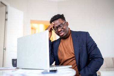 Stressed African man with headache looking at laptop screen in home office. Overworked Black businessman feeling exhausted, suffering from migraine, eye strain or financial stress during remote work.