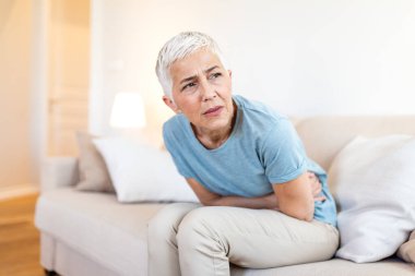 Yaşlı kadın koltukta karın ağrısı ve karın ağrısı çekiyor. Yetişkin bir kadın sindirim sorunu ya da evde regl sancısı çekiyor. Acı çeken yaşlılar için sağlık ve tıp konsepti.