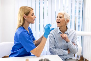 Mavi önlüklü profesyonel hemşire klinikte yaşlı bir kadının boğazını incelemek için tahta bir dil sıkacağı kullanıyor. Sağlık kontrolü, sağlık sigortası ve tanı konsepti. Doktor yaşlı hastaya danışmanlık yapıyor..