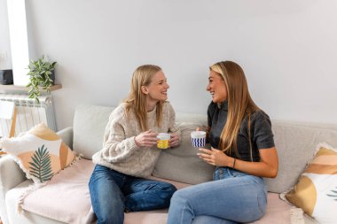 Kanepede gülüp kahve içen iki genç kadın. Arkadaşlık ve yaşam tarzı konsepti, oturma odasında sohbet eden mutlu bayan arkadaşlar. Modern apartmanda ortak ilgi alanları olan insanlar..