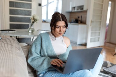 Mavi hırkalı genç bir kadın kanepede oturuyor ve dizüstü bilgisayarında daktilo yazıyor. Parlak, modern bir oturma odasında arka planda mutfağı olan serbest çalışan bir kadın. Yaşam tarzı portresi.