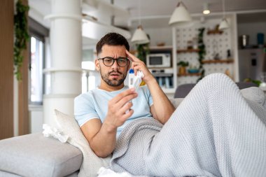 Hasta adam termometreyle ısıyı kontrol ediyor. Gözlüklü genç erkek battaniyenin altında kanepede yatıyor, ateşi, gribi ya da evde üşütmüş. Sağlık, ilaç ve virüs enfeksiyonu kavramı.